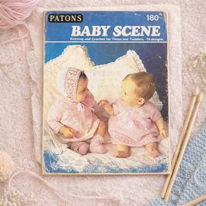 May include: A vintage book cover titled "BABY SCENE" by Patons, featuring two babies in pink knitted outfits. The cover is blue and white, with the text "Knitting and Crochet for Tinies and Toddlers - 15 designs". Pink yarn and knitting needles are also visible.