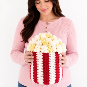 Peut inclure: Un panier en crochet en forme de pop-corn avec des rayures rouges et blanches, rempli de pop-corn blanc et jaune. Le panier est tenu par une personne portant un haut rose à manches longues et un jean bleu.