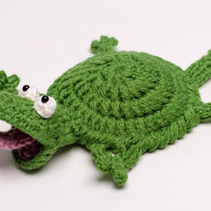 May include: A handmade green crochet alligator toy with white eyes and a pink tongue. The alligator has a textured, bumpy surface and is lying flat. The toy has four legs and a tail, all crocheted in the same green yarn.
