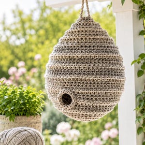 Op de afbeelding: Een handgemaakt vogelhuisje van gehaakt garen in beige en bruin tinten, met een ronde ingang, hangend aan een zwarte haak. Het hangt aan een witte houten balk. Een bol garen en een haaknaald liggen op een houten tafel.
