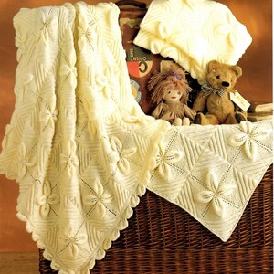 May include: Cream-colored knitted blanket with a floral pattern and scalloped edges. The blanket is draped over a wicker basket, alongside a matching pillow, a doll, and a teddy bear.