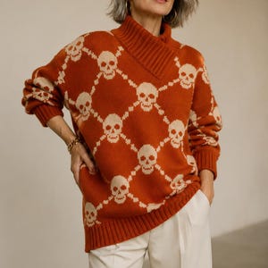 Puede incluir: Un suéter de punto color naranja óxido de manga larga con un patrón repetitivo de calaveras color crema dispuestas en una cuadrícula de diamantes. El suéter tiene un cuello alto doblado con detalles acanalados en el cuello, puños y dobladillo, junto a pantalones color crema.