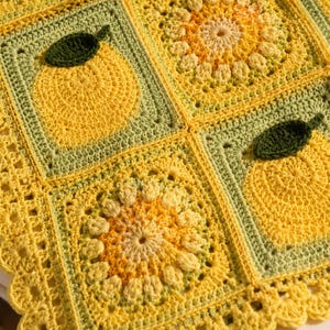 Könnte beinhalten: Eine handgefertigte Häkeldecke mit einem sich wiederholenden Muster aus gelben Zitrusfrüchten und grünen Blättern. Die Decke ist hauptsächlich gelb und grün, mit einem gewellten Rand. Das Fruchtmuster ist in quadratischen Blöcken zentriert.