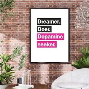 Op de afbeelding: Een ingelijste print met de tekst "Dreamer. Doer. Dopamine seeker." in zwarte en roze letters op een witte achtergrond. De print hangt aan een bakstenen muur, met planten en een moderne stoel op de voorgrond.