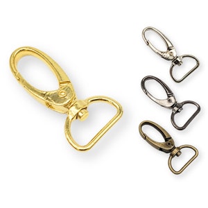 May include: A selection of metal swivel clasps in different finishes. The main clasp is gold-coloured, featuring a lobster claw design and a D-ring. Other clasps are silver, black, and bronze, all designed for crafting or accessory use.