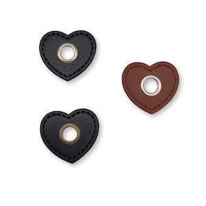 Può includere: Tre toppe di cuoio a forma di cuore con un occhiello di metallo al centro. Le toppe sono nere, marroni e marrone scuro.