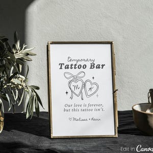 Könnte beinhalten: Ein gerahmtes Schild mit dem Text "temporary Tattoo Bar" und einem Herzdesign. Das Schild enthält auch den Text "Our love is forever, but this tattoo isn't." und die Namen Melissa & Kevin. Der Rahmen ist goldfarben.