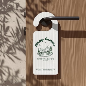Könnte beinhalten: Ein weißer Türhänger mit vertikaler Streifenstruktur und einer dunkelgrünen Grafik aus Bergen, Bäumen und einem See. Der Text lautet Happy Camper, MARGOT & SADIE'S ROOM sowie BETHANY'S BACHELORETTE, Joshua Tree, California in dunkelgrüner Farbe.