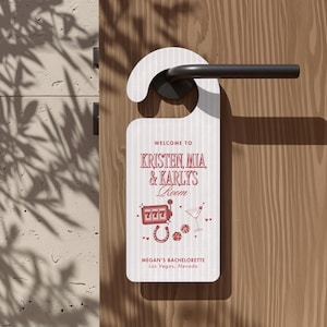 May include: A white door hanger with thin vertical stripes featuring red text that reads Welcome to Kristen, Mia, & Karly's Room and Megan's Bachelorette Las Vegas, Nevada. The design includes red icons of a slot machine, martini, and dice.