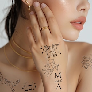 May include: A hand with temporary tattoos, including the text "& they lived happily ever after" and an illustration of Cupid. The hand also has a gold ring and is adorned with a gold necklace and earrings. The tattoos are black.