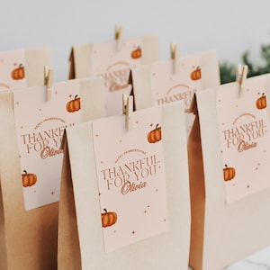 May include: Several brown paper gift bags with tags that read "Thankful for You" and a pumpkin graphic. The tags are attached with small wooden clothespins. The words "Happy Friendsgiving" are also on the tags.