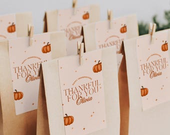 May include: Several brown paper gift bags with tags that read "Thankful for You" and a pumpkin graphic. The tags are attached with small wooden clothespins. The words "Happy Friendsgiving" are also on the tags.