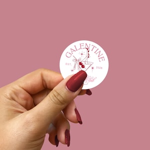 Könnte beinhalten: Ein weißer runder Aufkleber mit dem Text "GALENTINE Club" in burgunderfarbener Schrift. Das Design zeigt ein Martini-Glas mit Kirschen und Sternen. Der Aufkleber wird von einer Hand mit burgunderfarbenem Nagellack vor rosa Hintergrund gehalten.