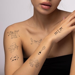 Puede incluir: Tatuajes temporales negros con diseños temáticos de boda. Incluye el texto "& they lived HAPPILY EVER AFTER", notas musicales, un cupido, palomas, una botella de champán, copas de champán e iniciales "K and R".