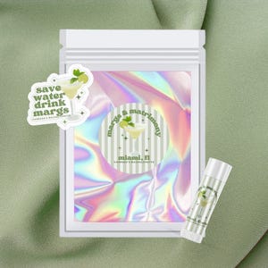 May include: A clear plastic bag with a white label that says "margs & matrimon" and "miami, fl" with a green and white striped background. The label also has a picture of a margarita with a lime wedge. There is a smaller sticker on the bag that says "save water drink margs" with a picture of a margarita. A lip balm with a similar design is also pictured.