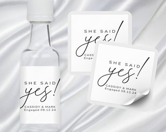 Pode incluir: Três adesivos quadrados brancos com texto preto que diz "SHE SAID yes!" com os nomes Cassidy & Mark e a data 09.12.24. Os adesivos estão em uma garrafa branca e uma superfície branca.