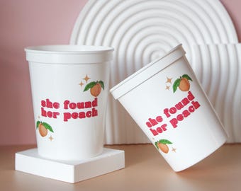 Può includere: Due bicchieri di plastica bianchi con la scritta "she found her peach" in rosso. Ogni bicchiere ha illustrazioni di pesche e foglie. Un bicchiere è in posizione verticale su un quadrato bianco, l'altro è inclinato.