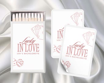 Può includere: Una scatola bianca di fiammiferi con un design rosa. La scatola ha il testo "Lucky in Love" e "Lexi's Bachelorette". Il design include carte da gioco e dadi.
