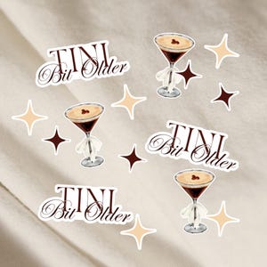 May include: A set of stickers featuring a cocktail glass with a brown drink and a white rim, and the text "TINI Bit Older" in a cursive font. The stickers are on a white background with small gold and brown stars.