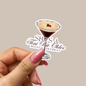 Könnte beinhalten: Ein Aufkleber mit einem Martini-Glas, gefüllt mit einer braunen Flüssigkeit und einer weißen Schaumkrone. Das Glas hat drei Kaffeebohnen obenauf. Der Text auf dem Aufkleber lautet "She's a Tini Bit Older" mit "Miley's Birthday" darunter.