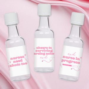 Puede incluir: Tres pequeñas botellas de vidrio con etiquetas blancas y texto rosa. Las etiquetas tienen diferentes mensajes relacionados con la escuela de enfermería. La primera etiqueta dice "nurses need shots too". La segunda etiqueta dice "cheers to surviving nursing school andi". La tercera etiqueta dice "andi nurse in progress LOADING..."