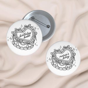 Może przedstawiać: Białe przypinki z czarno-białym wzorem tortu w kształcie serca. Tort jest ozdobiony kokardkami, perłami i napisem "birthday babe" lub "maria's bday". Przypinki mają metalową agrafkę z tyłu.