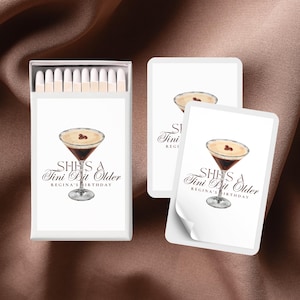May include: A set of white matchboxes with a white card attached to each. The card features a cocktail glass with a brown drink and a white topping. The text on the card reads "SHE'S A Tini Bit Older REGINA'S BIRTHDAY".