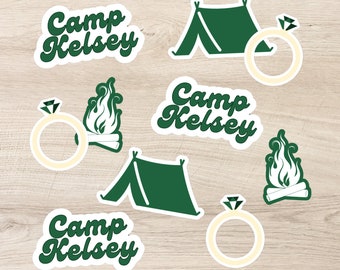 May include: A collection of stickers with a camping theme. The stickers include the text "Camp Kelsey" in a retro font, a green tent, a diamond ring, and a campfire. The stickers are on a light wood grain background.