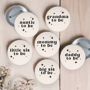 Op de afbeelding: Set van zes witte knopen met zwarte tekst en sterontwerpen. De knopen lezen "auntie to be", "grandma to be", "little sis to be", "mommy to be", "big sis to be" en "daddy to be".