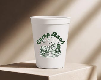 Puede incluir: Un vaso de plástico blanco con un estampado gráfico en verde oscuro que muestra un paisaje de montaña, un río, árboles, estrellas y una luna creciente. El texto "Camp Bach" aparece en verde oscuro con una tipografía curva y retro.