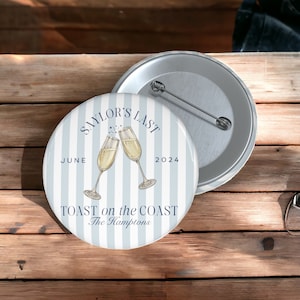 May include: A round button with a light blue and white striped background. The button features two champagne glasses clinking, with the text "Saylor's Last June 2024 Toast on the Coast The Hamptons."