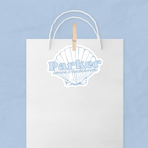 Puede incluir: Una concha azul con el texto "Parker carson's bachelorette" sobre un fondo blanco. La concha está unida a una bolsa de papel blanca con una pinza de madera.