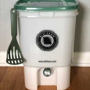 May include: White plastic Bokashi composting bucket with a green lid and a green scoop. The bucket has a tap at the bottom and a black and white logo that says "BLH Farm Grown in Missouri www.blhfarm.com"
