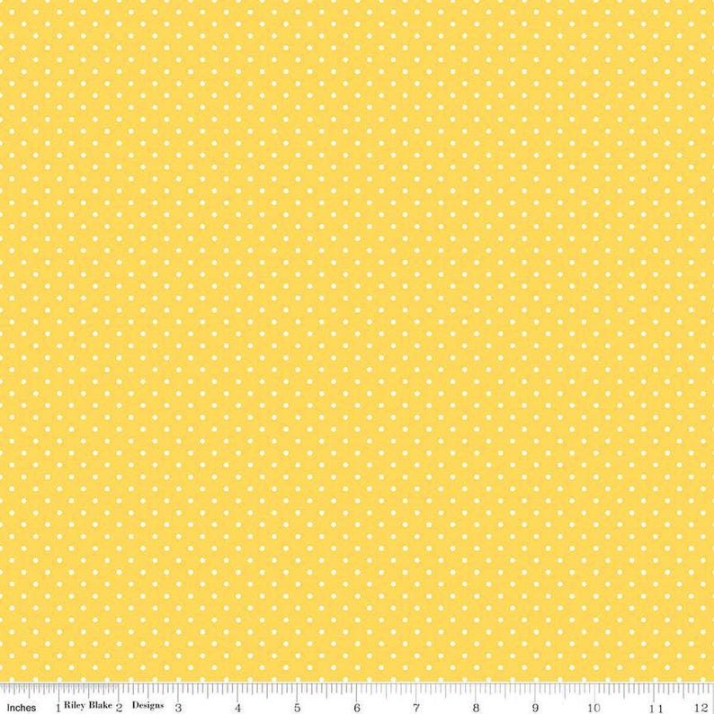 May include: A bright yellow fabric featuring a repeating pattern of small white polka dots. A white ruler is visible at the bottom edge, showing measurements in inches from 0 to 12, with the text Riley Blake Designs printed on it.