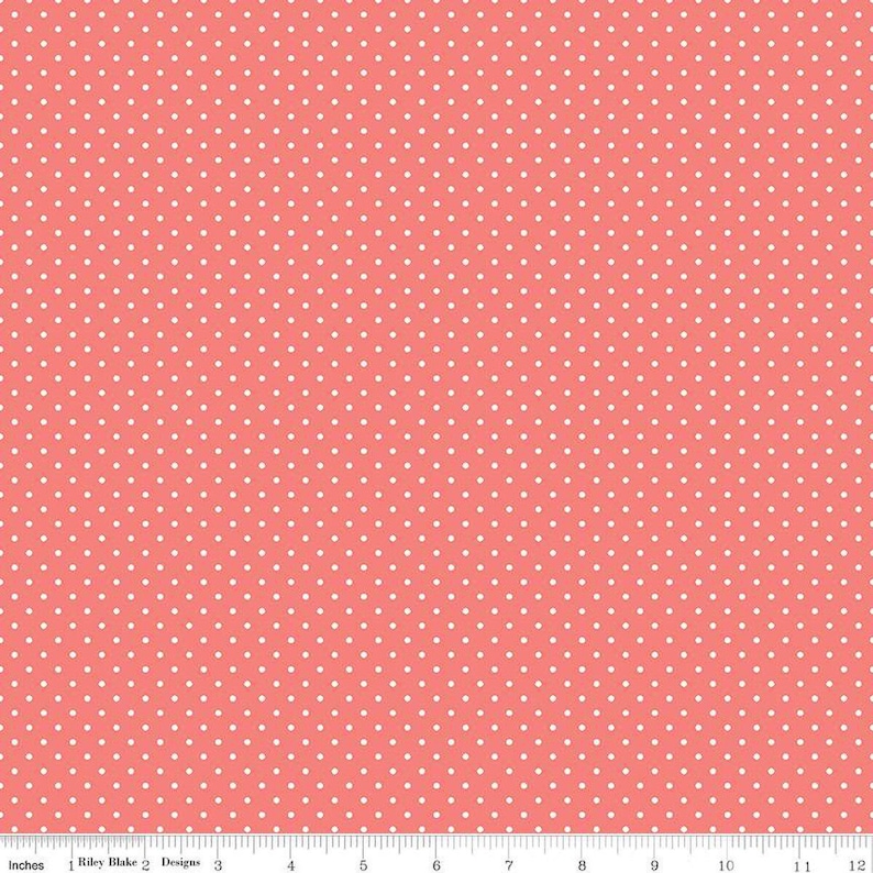 May include: A coral pink fabric featuring a repeating pattern of small white polka dots. A white ruler is visible along the bottom edge, showing a scale of 12 inches, with the text Riley Blake Designs printed near the left side.
