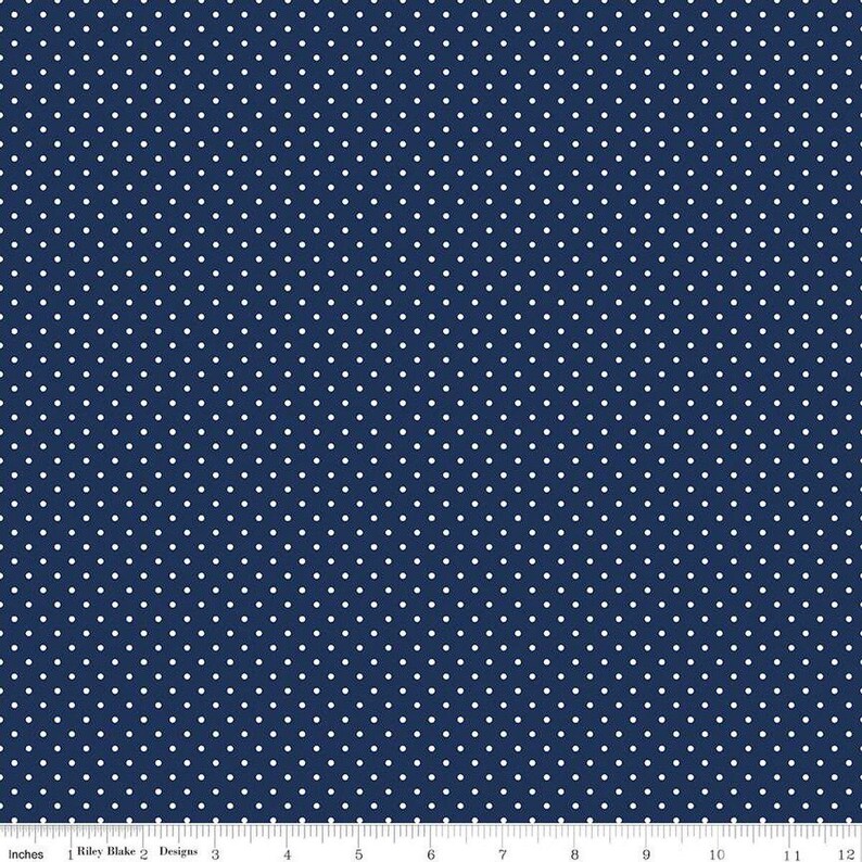 May include: A navy blue fabric featuring a repeating pattern of small white polka dots. A white ruler is visible at the bottom edge, showing a scale of 12 inches, with the text Riley Blake Designs printed along the measurement markings.
