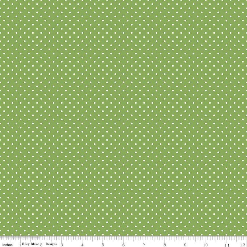 May include: A green fabric featuring a repeating pattern of small white polka dots. A ruler is visible at the bottom edge, showing a scale of 12 inches, with the text Riley Blake Designs printed along the measurement markings.
