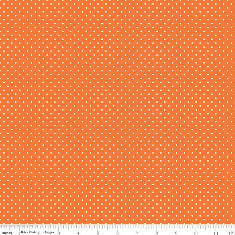 May include: A vibrant orange fabric featuring a repeating pattern of small white polka dots. A white ruler is visible at the bottom edge, showing a scale of 12 inches, with the text Riley Blake Designs printed near the left side.