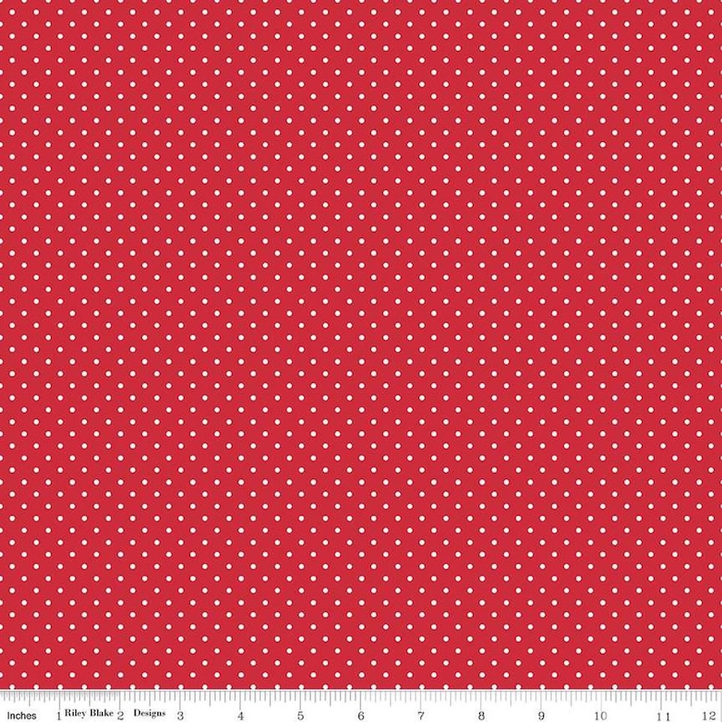 May include: A red fabric featuring a repeating pattern of small white polka dots. A white ruler is visible at the bottom edge, showing a scale of 12 inches, with the text Riley Blake Designs printed along the measurement markings.