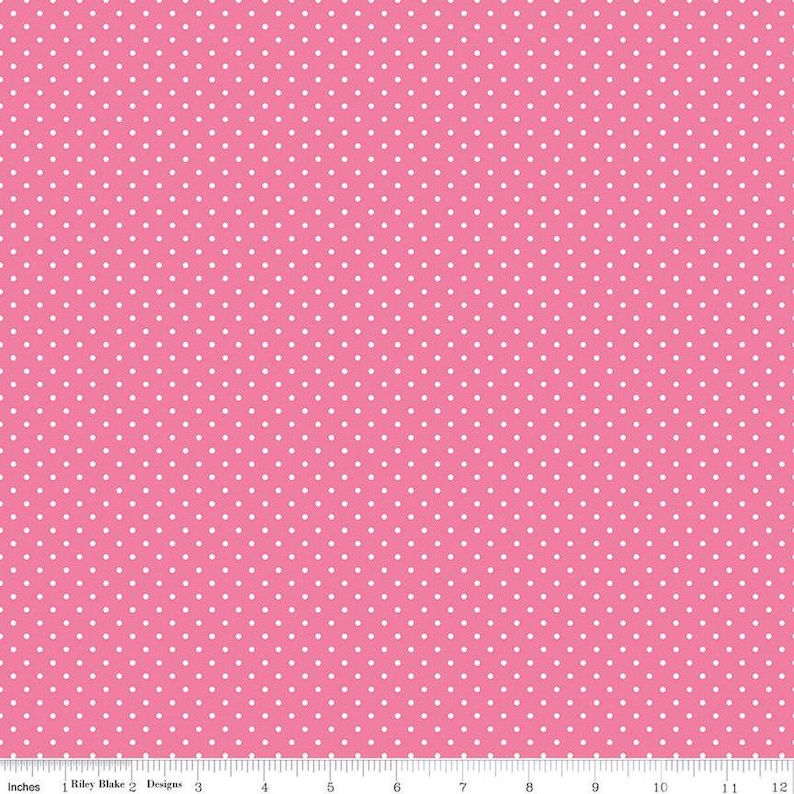 May include: A pink fabric featuring a repeating pattern of small white polka dots. At the bottom, a white ruler shows measurements in inches, with the text Riley Blake Designs printed clearly along the edge of the fabric.