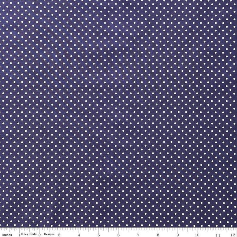 May include: A dark purple fabric featuring a repeating pattern of small white polka dots. A white ruler is visible at the bottom edge, showing a scale of 12 inches, with the text Riley Blake Designs printed near the left side.