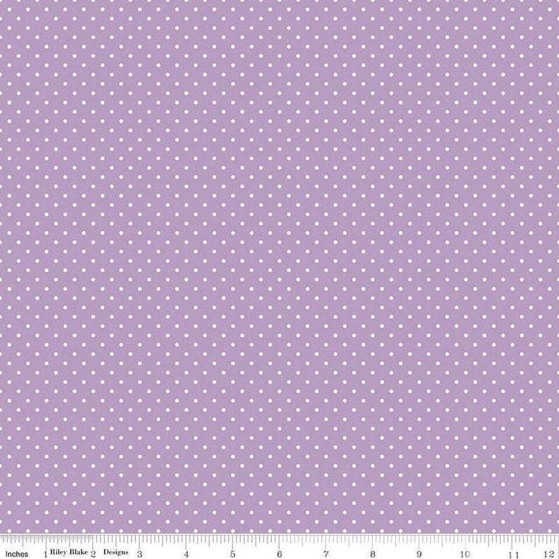 May include: A light purple fabric featuring a repeating pattern of small white polka dots. A ruler at the bottom edge shows a scale of 12 inches, with the text Riley Blake Designs printed near the one-inch mark.