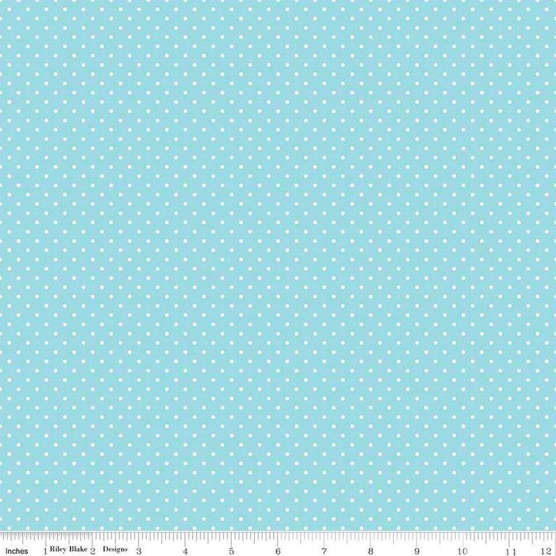 May include: A light blue fabric featuring a repeating pattern of small white polka dots. A white ruler is visible at the bottom edge, showing measurements in inches from 0 to 12, with the text Riley Blake Designs printed near the start.