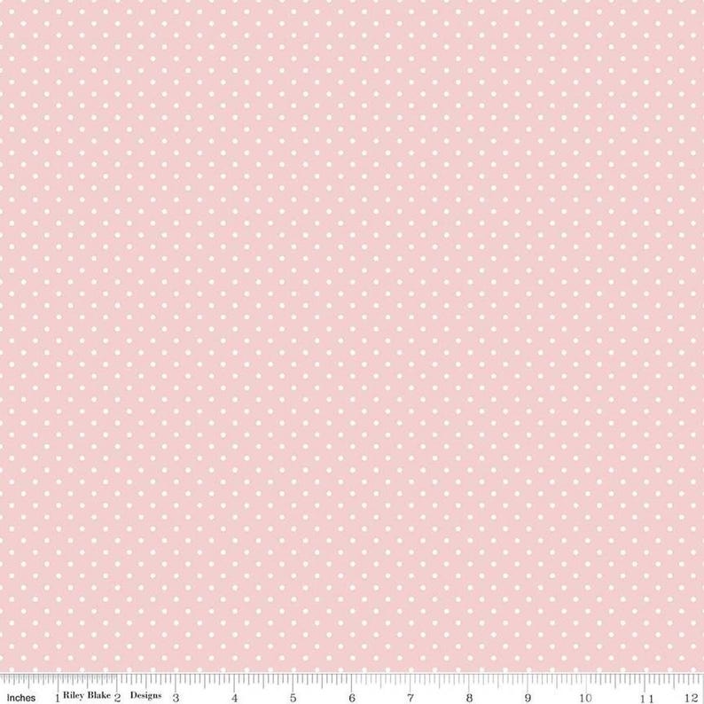 May include: A light pink fabric featuring a repeating pattern of small white polka dots. A white ruler is visible at the bottom edge, showing a scale of 12 inches, with the text Riley Blake Designs printed near the left side.