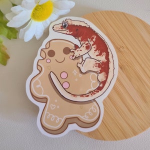 May include: A sticker featuring a cartoon gingerbread person with a red and white crested gecko on top. The gecko is curled around the gingerbread person's body. The sticker is on a light brown wooden surface.