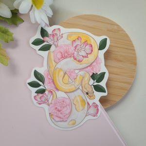 Può includere: Un adesivo raffigurante un serpente giallo e bianco attorcigliato tra fiori rosa e foglie verdi. Il serpente ha un motivo a squame dettagliato. L'adesivo è su una superficie rosa chiaro con un sottobicchiere di legno sullo sfondo.