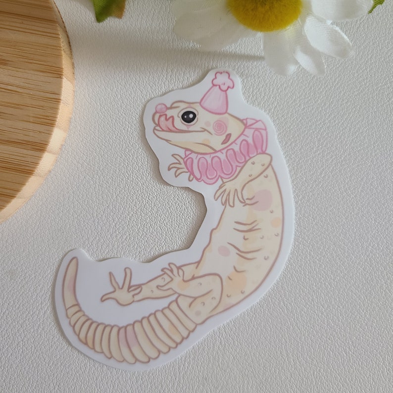 May include: A white gecko wearing a pink party hat and a pink ruffled collar. The gecko is drawn in a cartoon style with a light pink background.