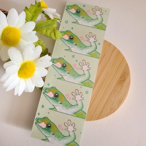 May include: A sheet of stickers featuring five green geckos with orange markings. Each gecko is waving with a pink heart above its head. The stickers are on a light green background with white stars.