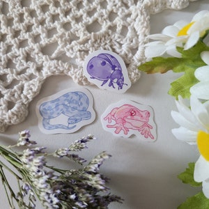 Könnte beinhalten: Drei Aquarell-Aufkleber mit einem lila Marienkäfer, einer blauen Schlange und einem rosa Frosch. Die Aufkleber befinden sich auf einem weißen Hintergrund mit einer gehäkelten Spitze und einem Lavendelstrauß.