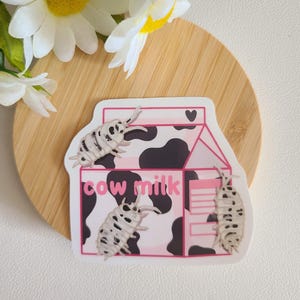 Op de afbeelding: Een witte sticker met een roze en zwart koe-print melkpak. Het pak staat "koemelk" en er kruipen drie witte pissebedden op.